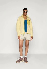 Inimene kannab beeži fliisi jope, sinise särgi üle, beeže lühikesi pükse, valgeid sokke, pruune jooksusuuski ja beeže prille, seistes ühetoonilise heledal taustal.