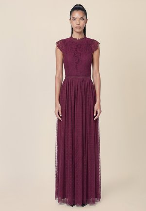 Woman stands facing forward wearing a sleeveless burgundy lace gown with a high neckline and a flowing skirt against a plain beige background.