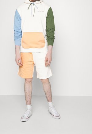 Felpa con cappuccio a sezioni blu, verde e crema; tasca arancione. Abbinata a pantaloncini arancioni e crema. Indossata con sneakers alte bianche.