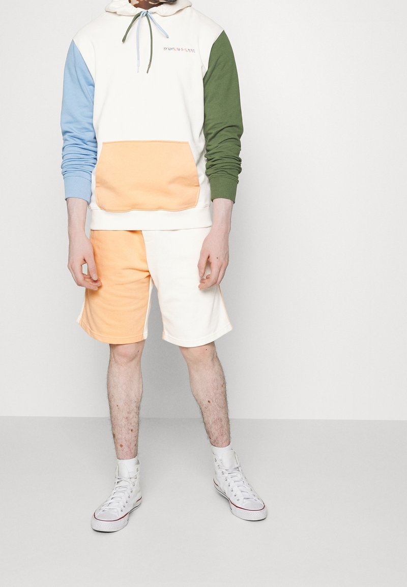 Huppari, jossa on sinisiä, vihreitä ja kermaisia osioita; oranssi tasku. Yhdistetty oransseihin ja kermaisiiin shortseihin. Päällä valkoiset korkeavartiset tennarit.