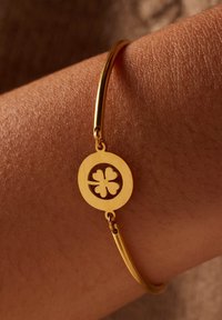 Gouden metalen bangle-armband met een ronde bedel met een uitgesneden klavertje vier ontwerp, gelegen op een gestructureerd huidoppervlak.