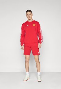 Rood sportieve sweatshirt en korte broek set gemaakt van stof. Kenmerkt zich door drie witte strepen op de mouwen en een logo op de borst. Inclusief witte schoenen.