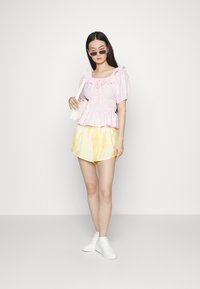 Top com mangas bufantes em padrões rosa e branco, com decote com babados, combinado com calções amarelos tie-dye e ténis brancos. Inclui uma bolsa minimalista.