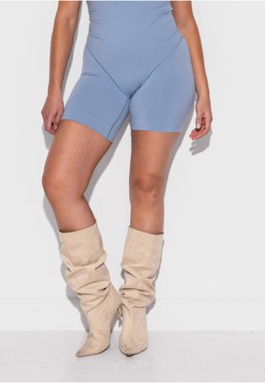 Frau trägt hellblaue eng anliegende Shorts und lässige beige Wildleder-Stiefel bis zur Mitte der Wade, steht vor einer weißen Wand.