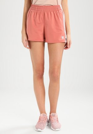 Kobieta w koralowych szortach Adidas z białymi bocznymi paskami i różowymi butami sportowymi, stojąca na jednolitym białym tle.