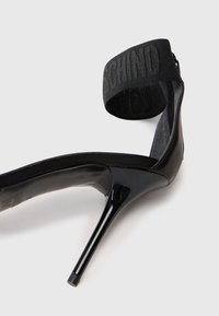 Μαύρο ψηλοτάκουνο παπούτσι με γυαλιστερό λούστρο, διαθέτοντας υφασμάτινη λωρίδα στον αστράγαλο με ανάγλυφο λογότυπο. Κομψός, μυτερός σχεδιασμός.