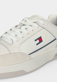Baskets basses blanches avec bout en daim, lacets blancs, panneau latéral perforé et logo Tommy Jeans en rouge, blanc et bleu sur le côté.