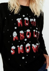 Musta neule, jossa on punaisista paljeteista kirjotut sanat "MITÄ SANOT LUMESTA" ja ylhäällä valkoisia paljettikuvioita, jotka muistuttavat lumesta.