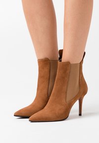 Ruskeat mokka-kangasta valmistetut kärjelliset nilkkasaappaat, joissa on elastiset sivupaneelit ja korkeat korot, käytössä paljaille jaloille valkoista taustaa vasten.