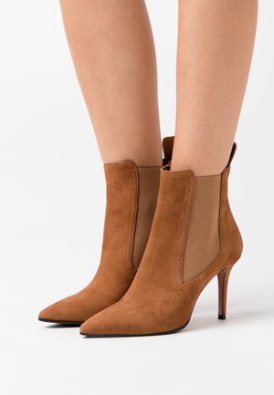 Bruine suede enkellaarsjes met puntige neus, elastische zijpanelen en stilettohakken, gedragen op blote benen tegen een witte achtergrond.