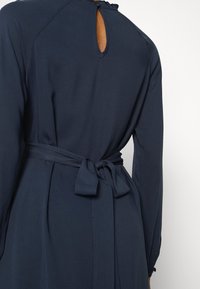 Mörkblå klänning med en nyckelhålsrygg, långa ärmar och ett knytbälte i midjan, tillverkad av ett slätt tyg med en flödande design.