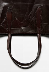 Brun läder toteväska med slät textur, med förlängda handtag och minimalistisk design. På ytan märks anmärkningsvärda veck.