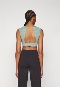 Šviesiai žalias sportinis liemenukas su tekstūruoto nėrinių dizainu, skylute nugaroje ir plačiu elastingu diržu su prekės ženklu.