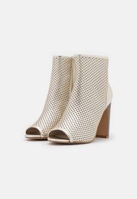 Zamszowe botki na wysokim obcasie z otwartym noskiem w metalizowanym odcieniu kremowym, zdobione perforowanym wzorem, gładkimi akcentami oraz smukłym drewnianym obcasem.