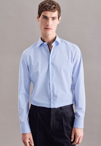 Ung mand iført en lyseblå langærmet skjorte med knapper og sorte bukser, stående mod en ensformig grå baggrund.