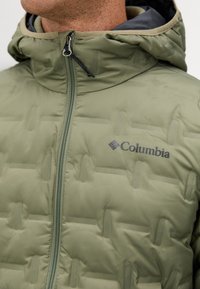 Maslinasto zeleni puffer jakna s kestenjastim dizajnom, koja ima prednji zatvarač, kapuljaču i crni Columbia logo na lijevoj strani.
