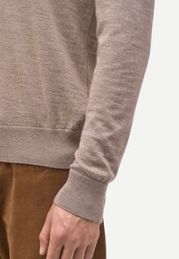 Beiger Strickpullover, weiche Textur, gerippte Bündchen und Saum. Der Ärmel reicht bis zur Mitte des Unterarms, kombiniert mit braunen Cordhosen.