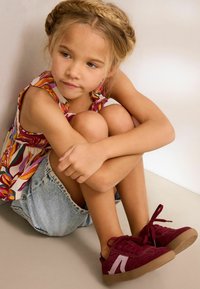 Baskets en suède bordeaux avec une semelle en caoutchouc texturé et un accent blanc, associées à un haut fleuri et à un short en denim.