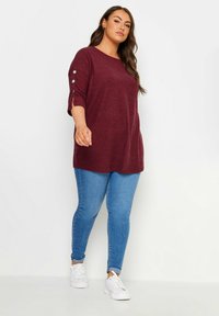Burgundy tunika dengan lengan tiga perempat, menampilkan aksen kancing pada lengan, dipadukan dengan celana skinny biru muda dan sneaker putih.