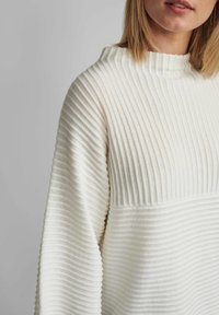 Sweter w kolorze kremowym z pionowym prążkowym wzorem, z wysokim kołnierzem i szerokimi rękawami. Szew oddziela teksturowane i gładkie sekcje.