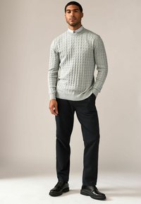 Pull en tricot gris avec un col haut, associé à un pantalon foncé et des chaussures noires. Texture lisse et détail de design texturé.