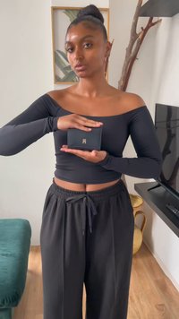 Portefeuille en simili cuir noir avec un logo doré, tenu dans la main d'une personne. L'arrière-plan présente un canapé vert et un sol en bois clair.