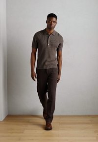 Camisa polo marrón de manga corta con un patrón texturizado y tapeta de tres botones, combinada con pantalones marrones oscuros y zapatos marrones.