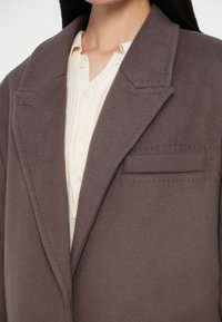 Bruine wolmix jas met een gesneden kraag, één zak en subtiele stiksels. Draagt over een lichtgekleurde, met knopen sluitende shirt.