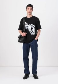 Juodas marškinėliai su grandinės grafika, mėlyni džinsai ir juodi kulkšninių botai. Modelis laiko mažą juodą per petį nešiojamą krepšį. Paprastas ir modernus dizainas.