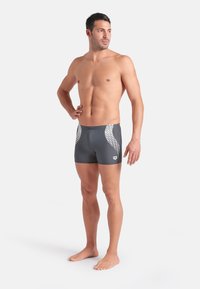 Costumi da bagno da uomo in grigio scuro con accenti geometrici bianchi sui lati. Realizzati in materiale liscio e elastico. Lunghezza fino a metà coscia.
