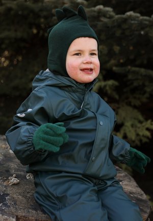 Tuta impermeabile verde con texture morbida, dotata di cappuccio e cappello di maglia verde con punte. Bambino con guanti abbinati, seduto su un tronco.