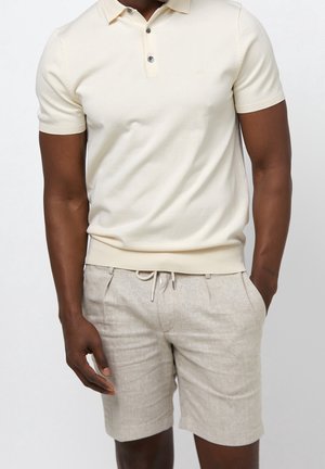 Homme portant un polo à manches courtes crème et un short beige clair avec cordon, debout avec une main dans la poche sur fond blanc.