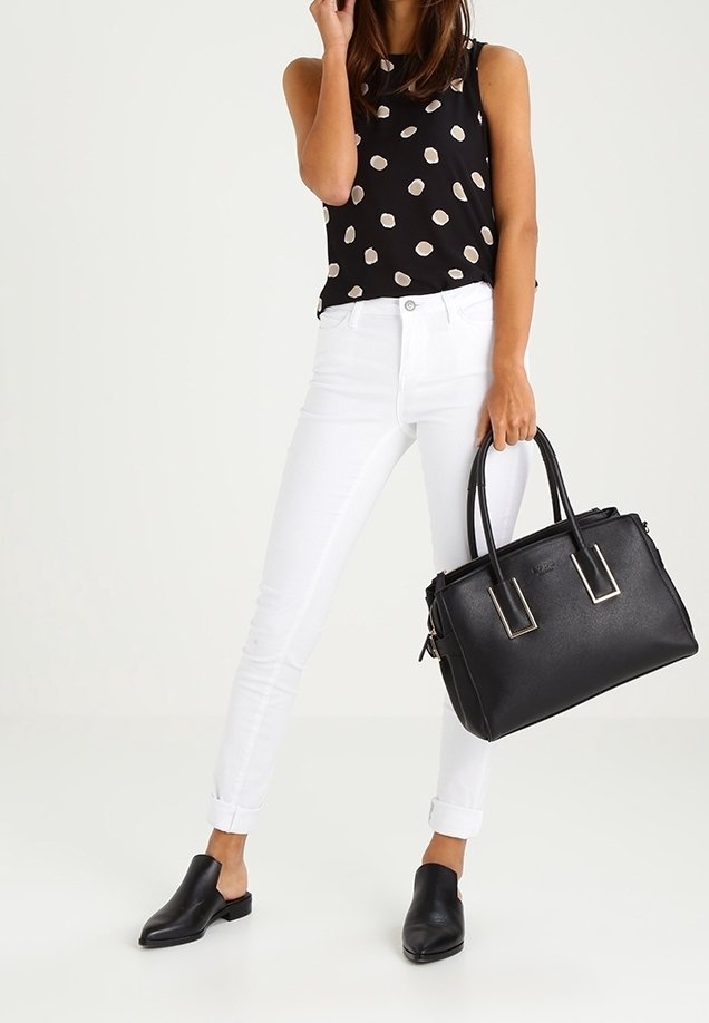 Femme portant un haut noir à pois, pantalon blanc roulé, chaussures noires sans lacets, tenant un grand sac à main noir par ses poignées.