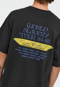 Sort bomulds T-shirt med et grafisk design i blå og gul tekst, der viser "WORLD SLAVERY TOUR 84-85" over et ørnedesign.