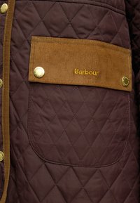Tissu matelassé marron avec rabat de poche en velours côtelé beige, fixé par deux boutons dorés et broderie jaune avec le logo "Barbour".