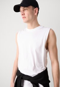 Camisa blanca de algodón sin mangas con cuello redondo, acompañada de un suéter negro atado a la cintura. Presenta un fondo neutro.