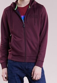 Sudadera con cremallera burdeos con acentos verdes, fabricada en un tejido suave, con puños y dobladillo acanalados, que presenta un logotipo en el pecho y bolsillos laterales.