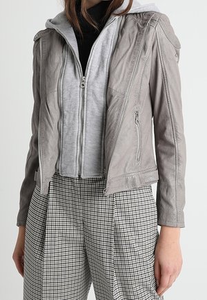 Femme portant des vêtements superposés comprenant une veste en cuir grise sur un sweat à capuche zippé gris clair et un pantalon à carreaux noir et blanc.