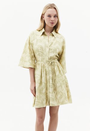 Młoda kobieta w jasnosłomkowej sukience w kwiaty z guzikami, szerokimi rękawami i paskiem do wiązania, stojąca na białym tle.