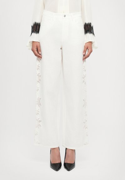 White wide-leg pants featuring lace detailing along the sides, complemented by a black blouse and pointed black heels.