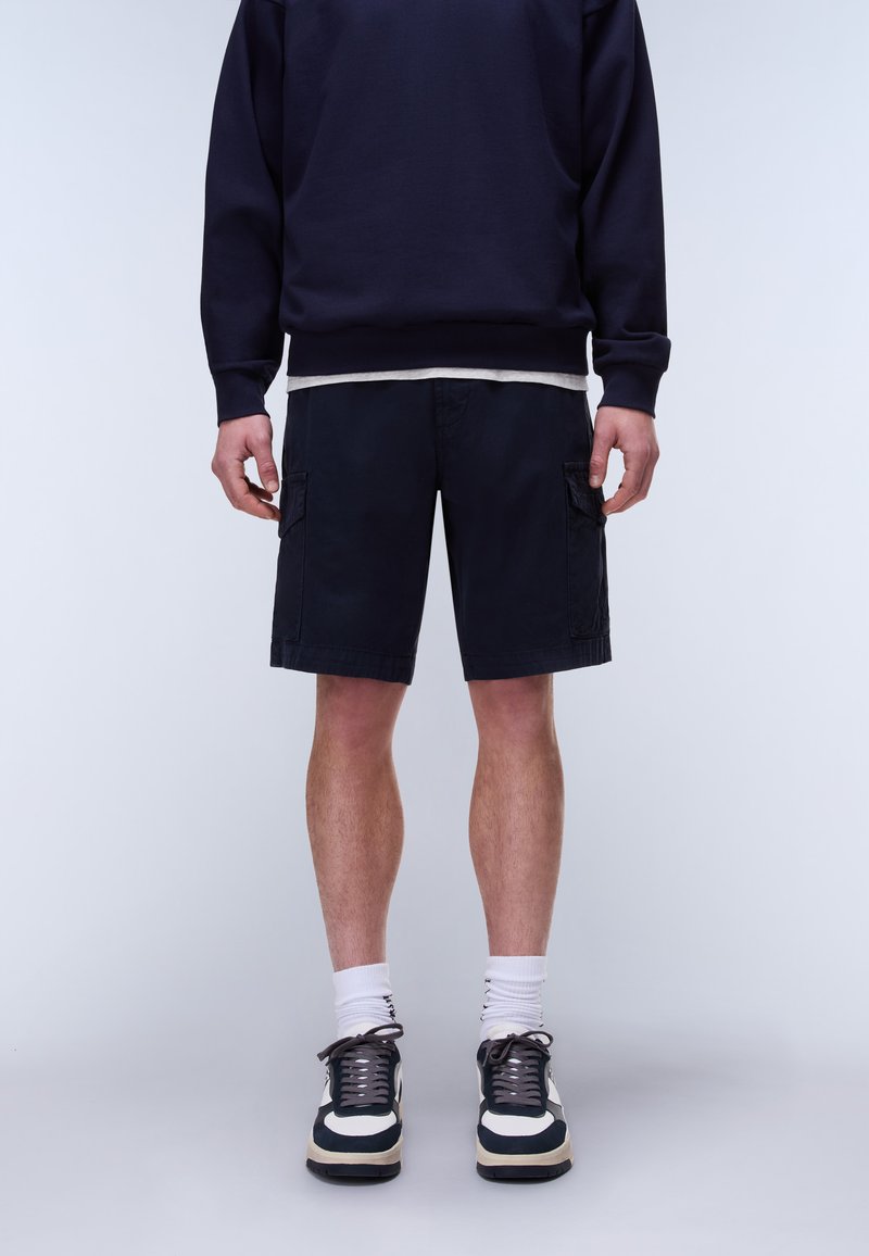 Mann trägt dunkelblaue Cargo-Shorts, einen marineblauen Pullover, weiße Socken und schwarz-weiße Sneakers vor einem hellgrauen Hintergrund.