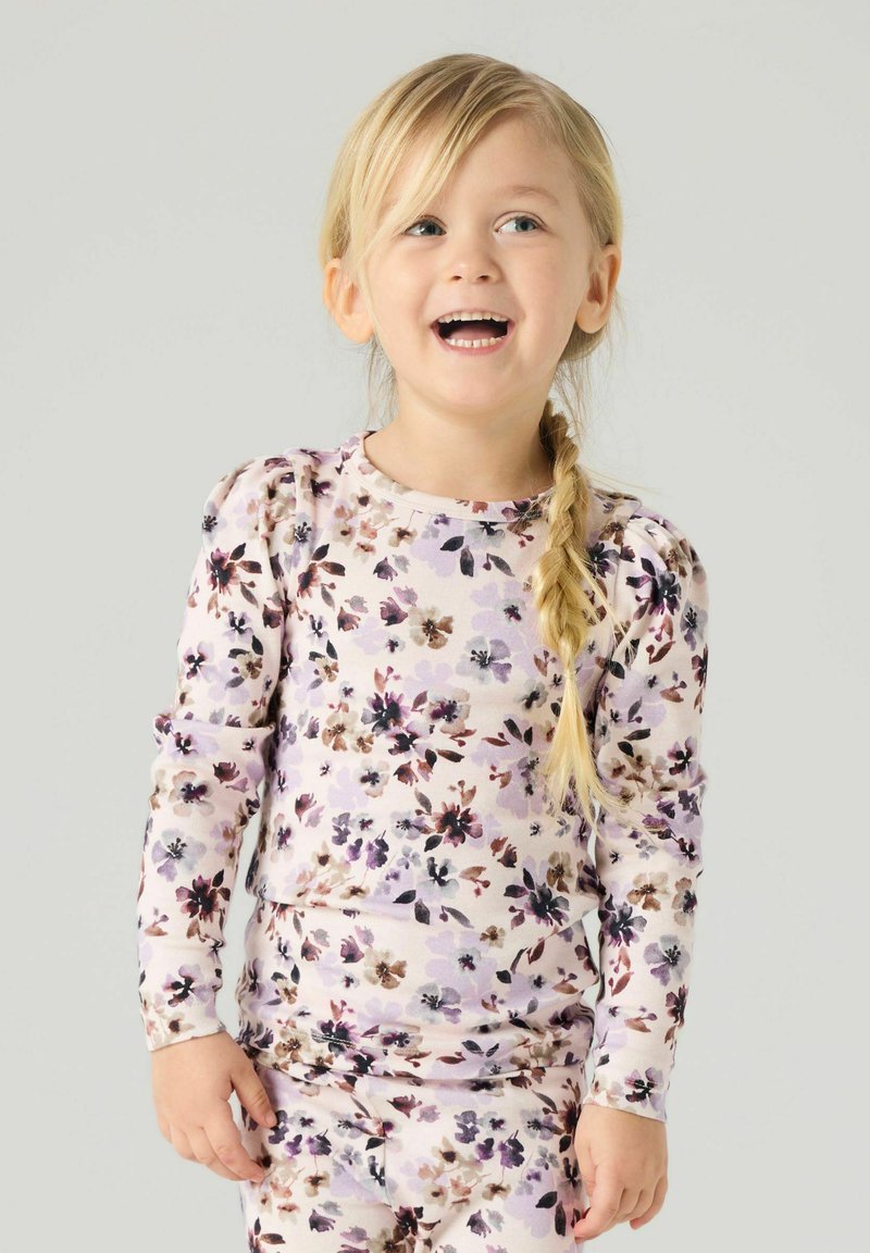 Jeune fille avec une tresse blonde souriante, portant une chemise à manches longues à fleurs violet clair et un pantalon assorti sur un fond uni.