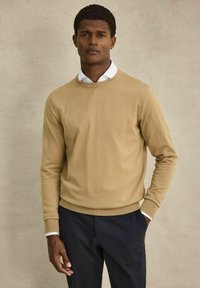 Beige neulepaita, joka on valmistettu sileästä neuleesta, jossa on ribbireunat hihansuissa ja helma. Paita on puettu valkoisen kauluspaidan ja tummien housujen yli.