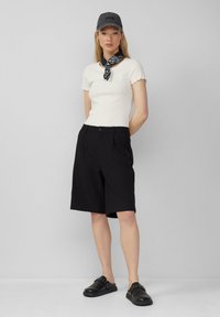 Weißes geripptes kurzärmliges Oberteil, schwarze maßgeschneiderte Shorts, schwarze Kappe und schwarze Slipper. Ein gemusterter Schal ist um den Hals gebunden.