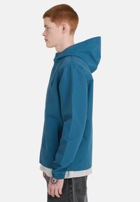 Persona joven con cabello rojo, vestida con una chaqueta con capucha color verde azulado, camiseta gris claro y jeans gris oscuro, de pie de lado contra un fondo blanco.