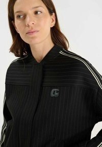 Svart jacka med smalrand och ståkrage, med vita accentlinjer och en logo på framsidan. Materialet verkar slätt och strukturerat.