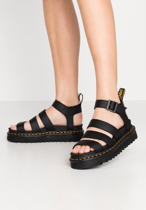 Benedenbenen met zwarte leren platformsandalen met drie banden en een gesp, met gele stiksels op dikke zolen.