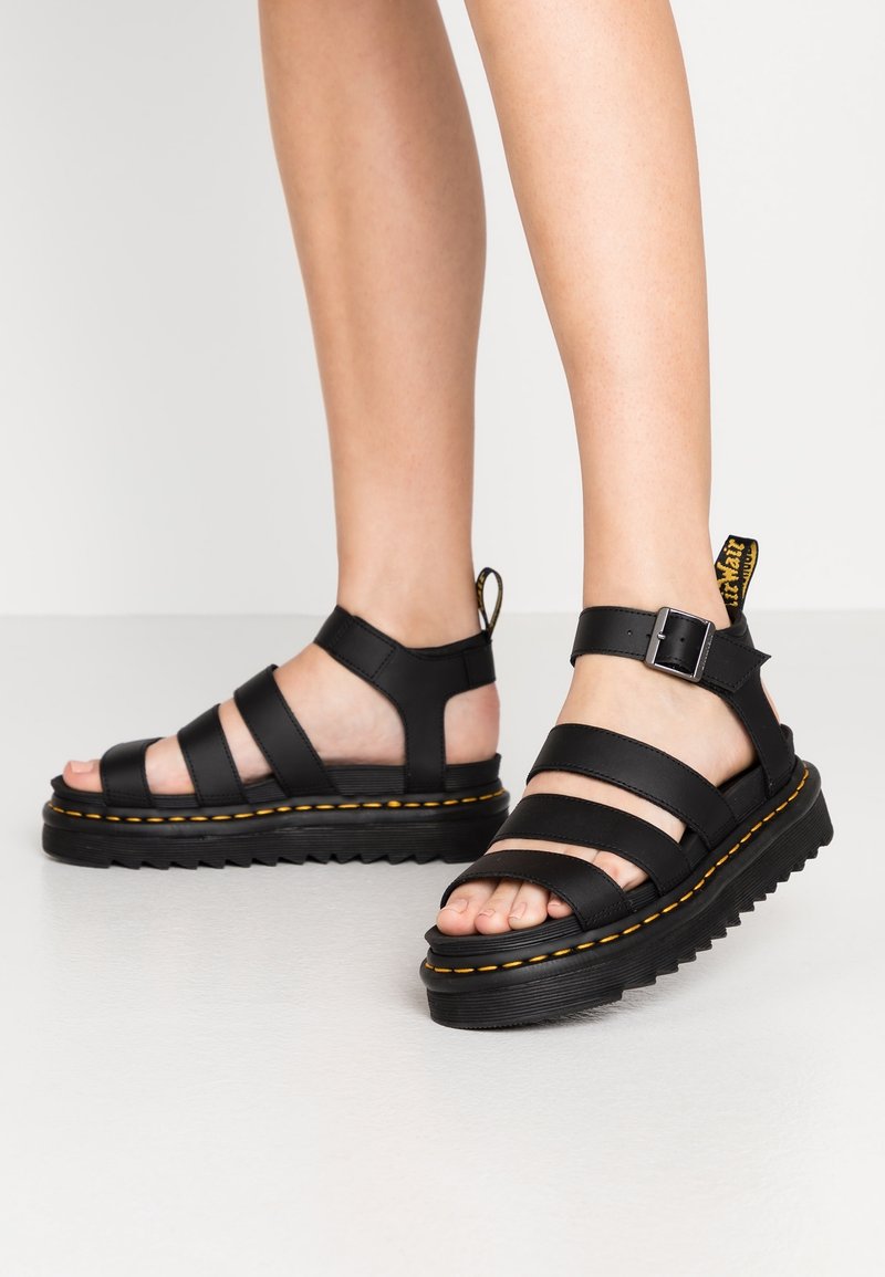 Jambes portant des sandales plateformes en cuir noir avec trois brides et une boucle, avec des coutures jaunes sur des semelles épaisses.