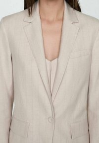 Veste de costume beige cintrée à rayures fines, fermeture à un bouton et revers crantés ; dotée de deux poches avant et d'une texture lisse.