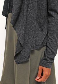 Tmavě šedý top s dlouhým rukávem a volným střihem v kombinaci s olivově zelenou sukní, ukazující pohodlný vzhled s rukou odpočívající podél těla.